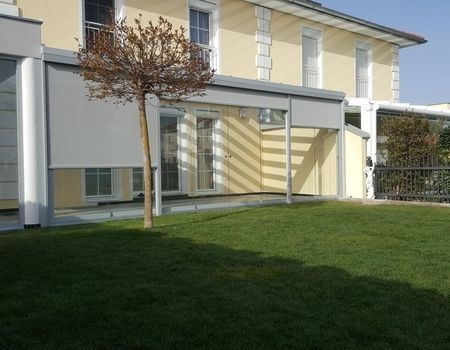 Eine Terrasse mit einem Glasdach und Glasschiebetüren im Vordergrund steht ein Baum
