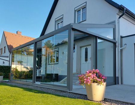 Ein großer Sommergarten mit Glasschiebetüren  auf einer großen Terrasse. Im Vordergrund steht ein Blumentopf mit bunten Blumen